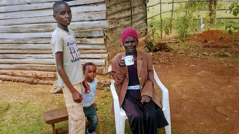 A woman sits on a chair in front of her home with two grandchildren at her side.
