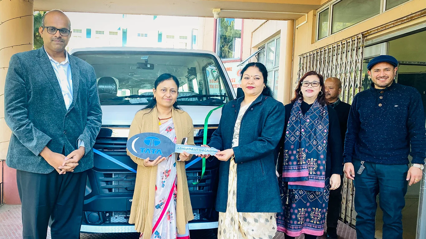 A woman hands over a large key to another woman. They stand in front of a van