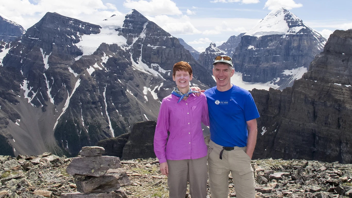 A man and woman stand together on a mountain pass. There are snowcapped peaks in the background.