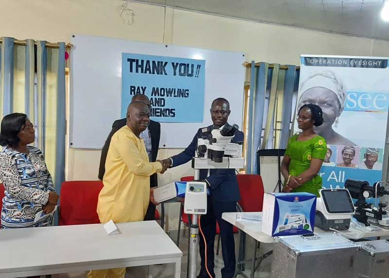 Two men shakes hands as an eye care equipment is exchanged. The poster behind them says "Thank you Ray Mowling and friends"