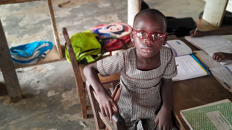 A girl wearing glasses and a school uniform sits at a desk.