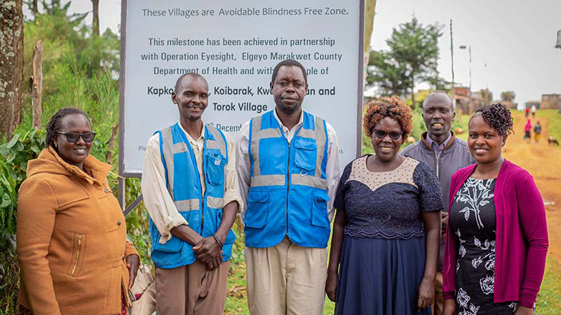 Three women and three men pose in front of a large poster announcing an Avoidable Blindness-Free Zone in a rural setting in Elgeyo Marakwet