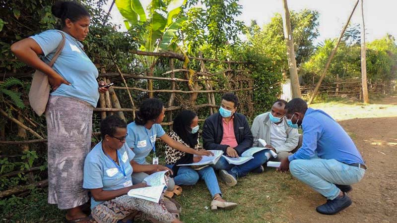 : A group of community health workers wearing matching blue t-shirts meet with Yashwant Sinha and some program managers in the shade beneath a fence in a rural setting in Bahir Dar town