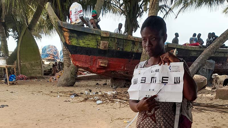 A woman holds up an eye chart to people to people not seen in the photo. Behind her