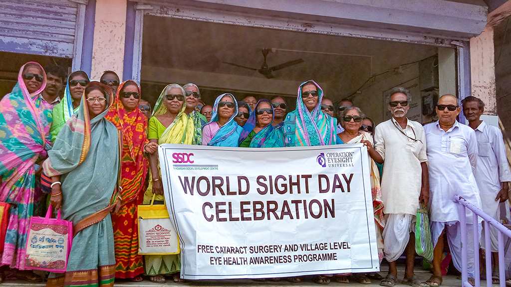 A group of people stand outside of a building holding a sign that reads "World Sight Day Celebration". All wear big