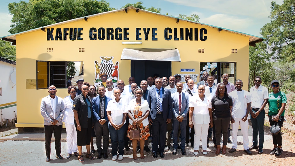 A group of people stand in front of a small yellow building with the words "Kafue Gorge Eye Clinic" painted across the front.|||