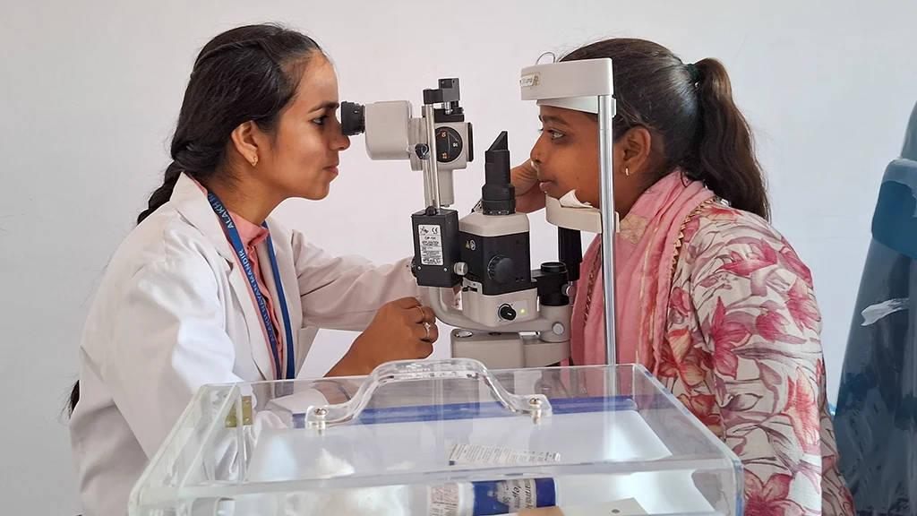 A woman in a lab coat examines the eyes of another woman through a slit lamp.|||||A woman holds up an eye chart. She is outdoors
