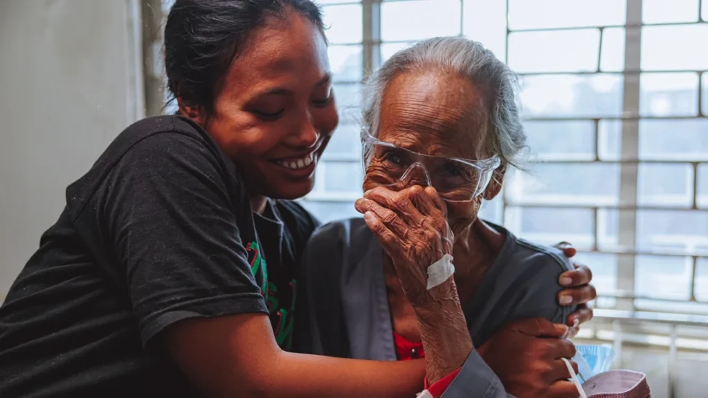 A young woman hugs a white-haired woman. The older woman wears a hospital gown and protective