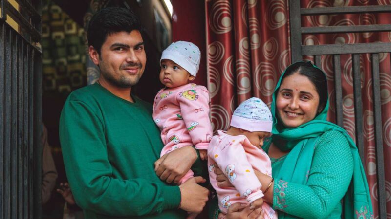 A man and his wife stand holding twin baby girls dressed identically in pink onesies.|Family smiling while carrying twin girls in Moradabad