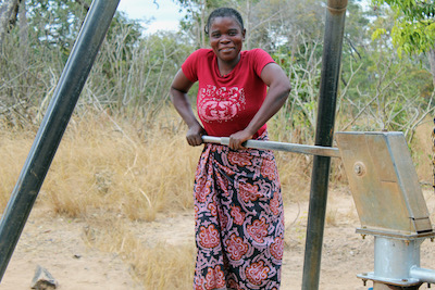A woman collects water from a borehole and smiles at the camera|A woman smiles as she carries water in a yellow bucket