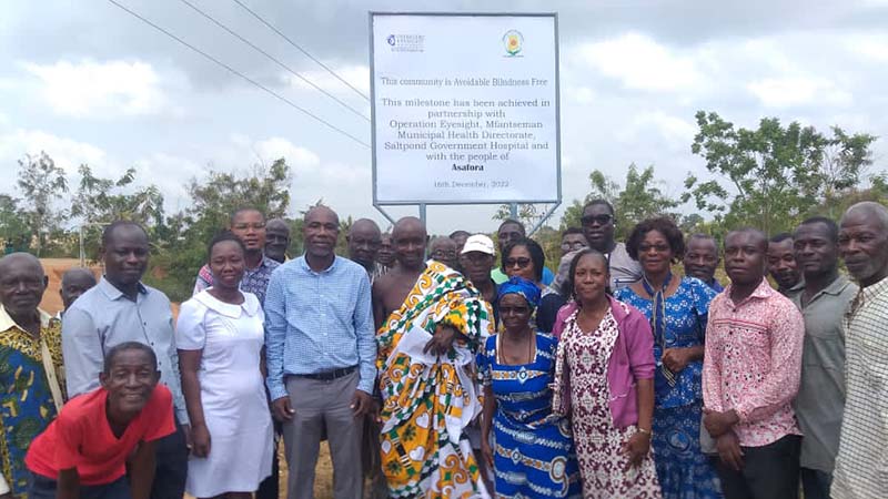 Community members and dignitaries stand in front of an Avoidable Blindness-Free village billboard.|Community members and dignitaries gather in front of an 'Avoidable Blindness-Free Village' sign.