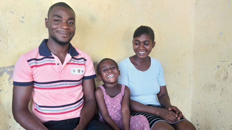 A young girl sits in between her parents smiling at the camera|Blessing smiles as she stands next to her mother|A curved black metallic object|A little girl