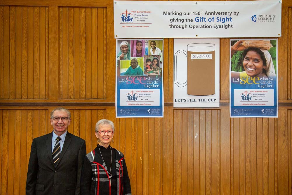 A senior woman and man post for a photo in front of a few posters that show a fundraising campaign for Operation Eyesight and its progress. The text on one of the posters reads "Making our 150th anniversary by giving the gift of sight through Operation Eyesight."|The 150-year-old First Baptist Church in Petrolia