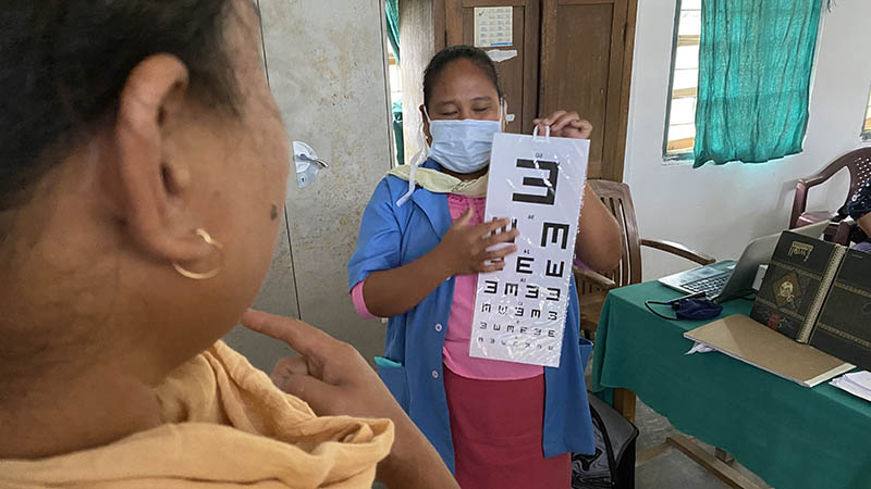 A female health worker holds up an eye chart for a female patient.|A female health worker holds up an eye chart in front of a female patient.