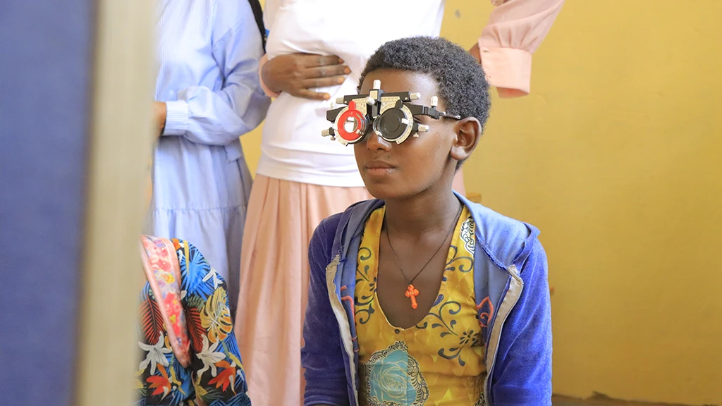 A girl wears a pair of refracting frames while getting an eye exam.|Two pointed city towers are lit up in blue and orange lighting.|A group of women and hcildren sit in a waiting room.|A group of men and women in matching white polo shirts hold up a large banner that reads "Affordable