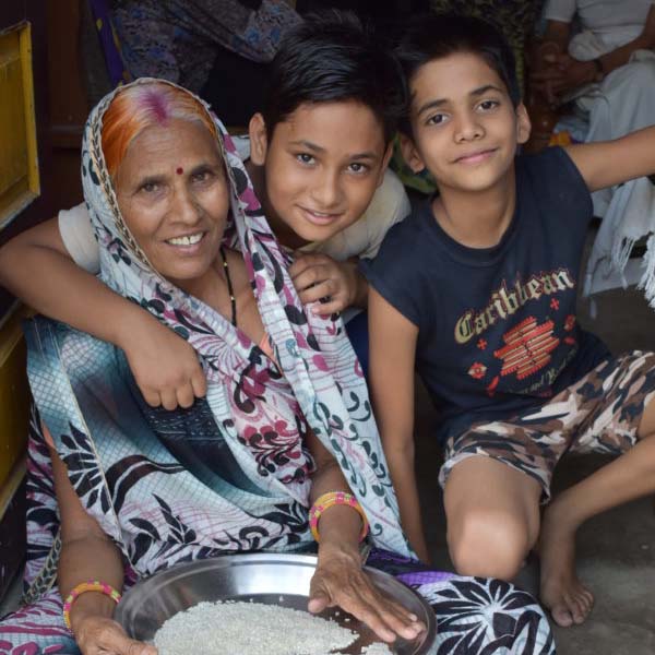 Photo of a woman sitting with two young boys|Premabai sits with her grandson as she sorts through a plate of rice grains|Premabai taking care of grandchild