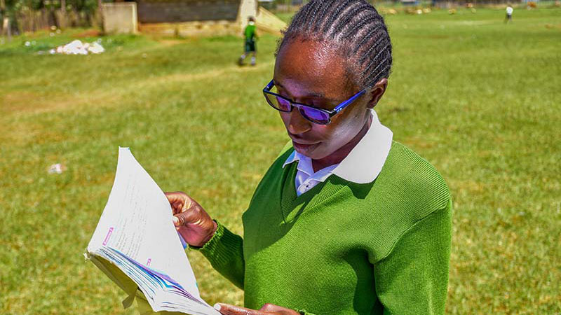 A teenaged girl in a green sweater and blue eyeglasses stands reading a book in a schoolyard.|A teenaged girl in a green sweater and blue eyeglasses stands with her peers|A teenaged girl in a green sweater and blue eyeglasses stands reading a book in a schoolyard.